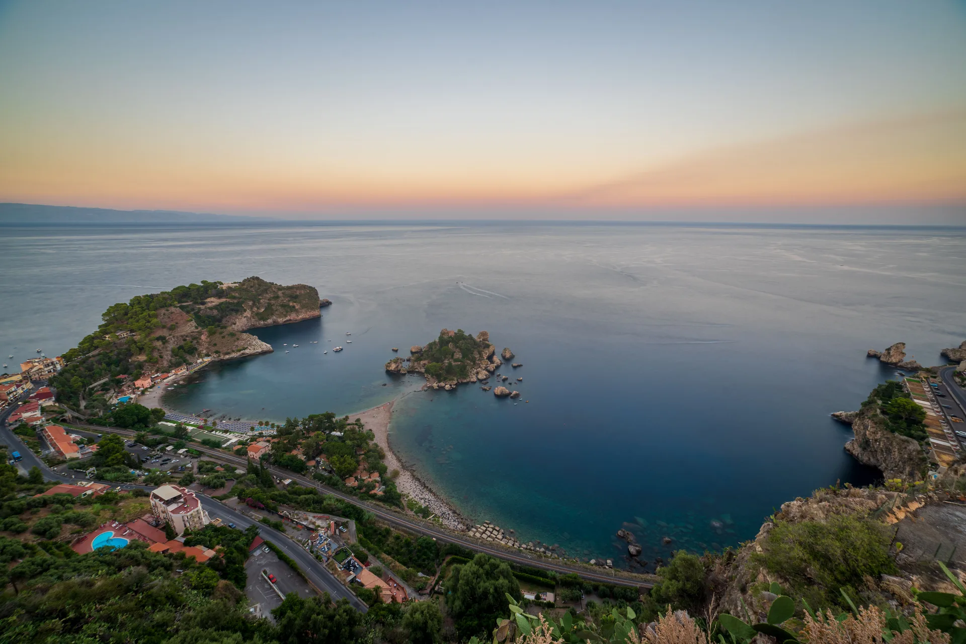 Elevated dusk view of Isola Bella near Taormina, a small pine-covered island connected to the coast by a sandbar, glowing water reflecting the sunset sky.